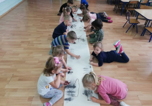 Dzieci siedzą na podłodze wzdłuż rozłożonego z rolki kartonu po obu jego stronach i rozpoczynają rysowanie.